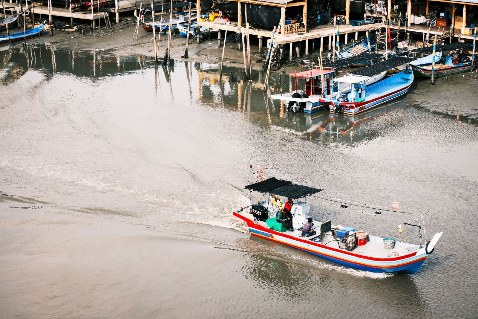 A colorful fishing boat is moving along a calm river near stilt houses in Perlis, Malaysia.