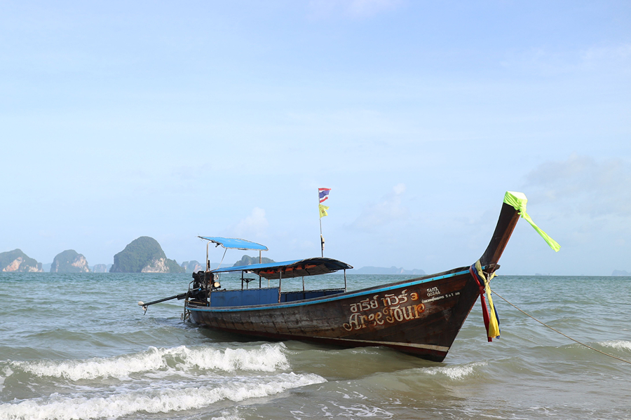 A traditional Thai longtail boat on a quiet beach, with islands in the background under a clear blue sky