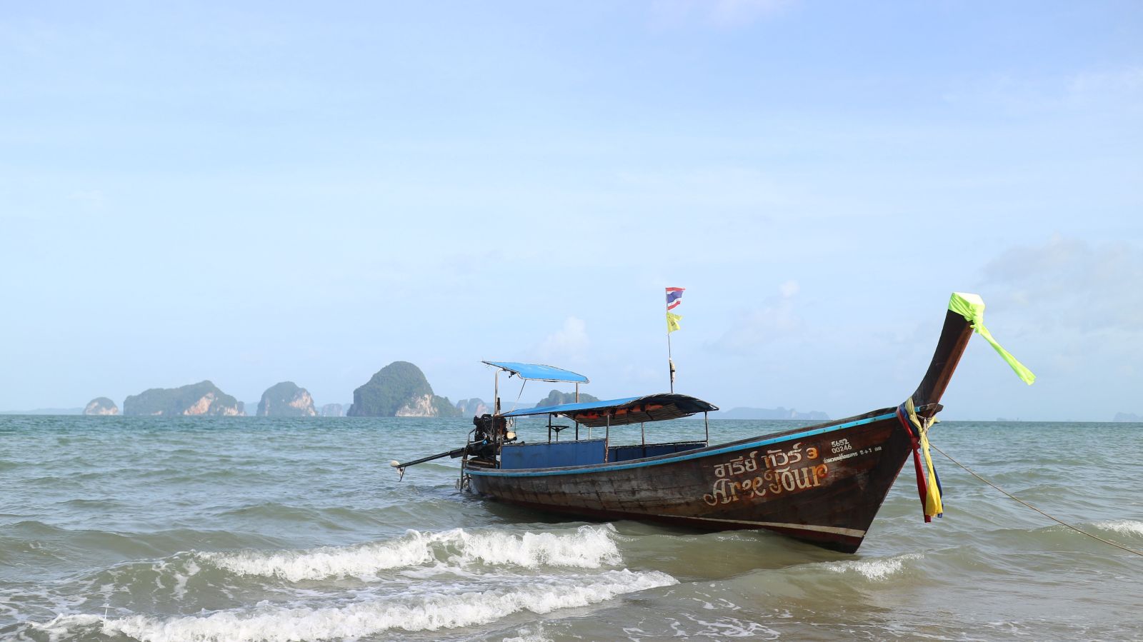 A traditional Thai longtail boat on a quiet beach, with islands in the background under a clear blue sky.