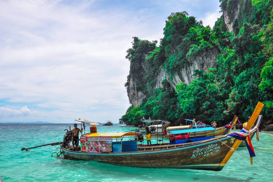 A traditional longtail boat is anchored in turquoise waters near limestone cliffs in Phang Nga, Thailand.