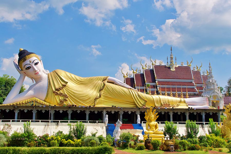 The gold-plated reclining Buddha statue stands out against the clear blue sky within the grounds of a stunning Thai temple.